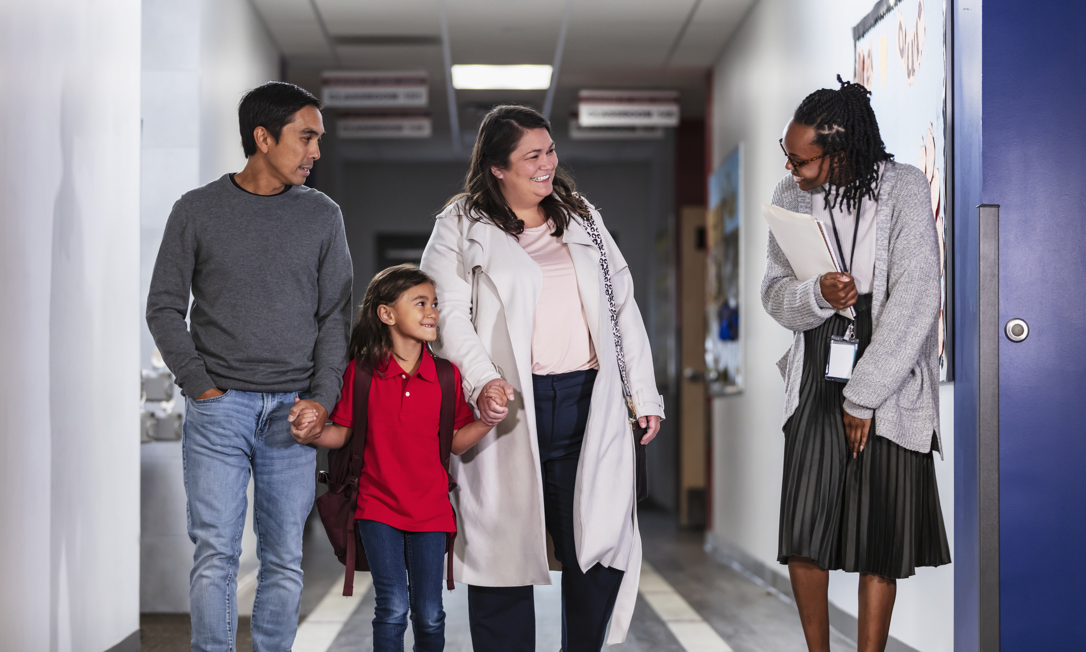 Two parents bringing their daughter to school, talking with an elementary school teacher or principal in the hallway
